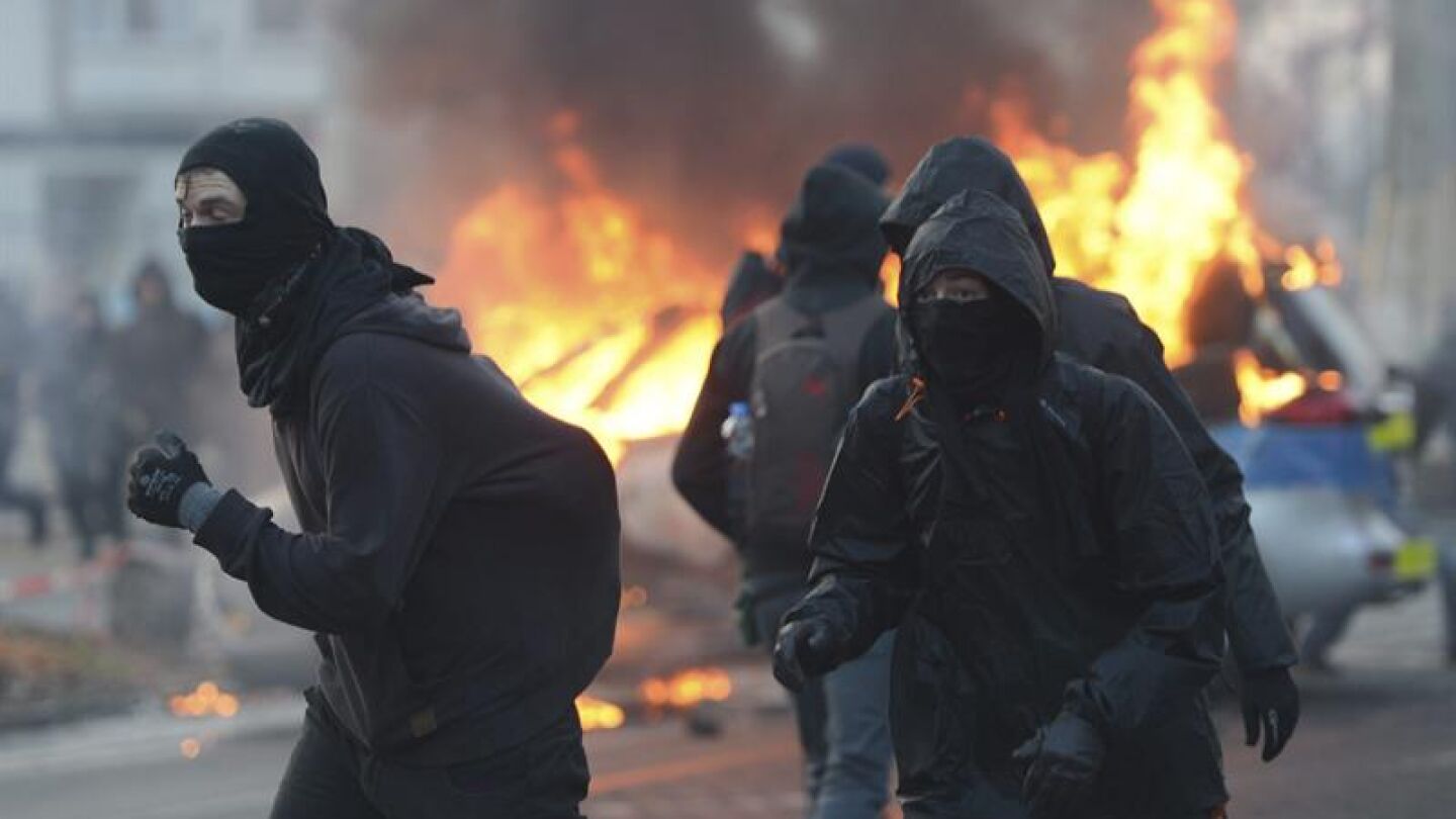 Manifestantes se cubren el rostro junto a un veh&iacute;culo policial que arde ante la sede del BCE
