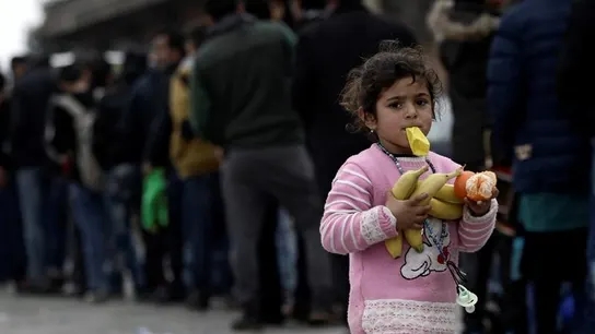 Una niña camina con fruta en las manos mientras muchos otros refugiados esperan comida Una niña camina con fruta en las manos mientras muchos otros refugiados esperan comida