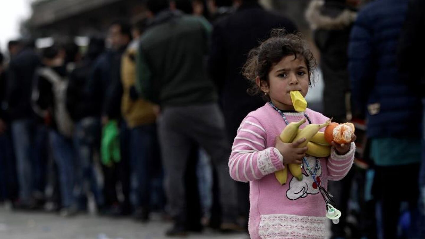 Una ni&ntilde;a camina con fruta en las manos mientras muchos otros refugiados esperan comida