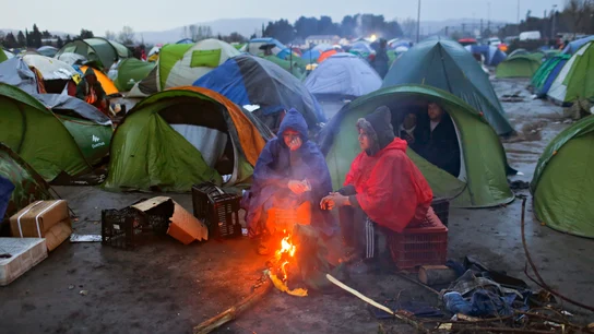 Varios refugiados se calientan junto al fuego entre sus tiendas de campaña en un campamento de refugiados Varios refugiados se calientan junto al fuego entre sus tiendas de campaña en un campamento de refugiados