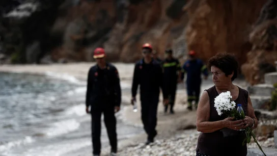 Varios bomberos buscan a desaparecidos mientras una mujer sostiene un ramo de flores tras el incendio forestal de Mati Varios bomberos buscan a desaparecidos mientras una mujer sostiene un ramo de flores tras el incendio forestal de Mati