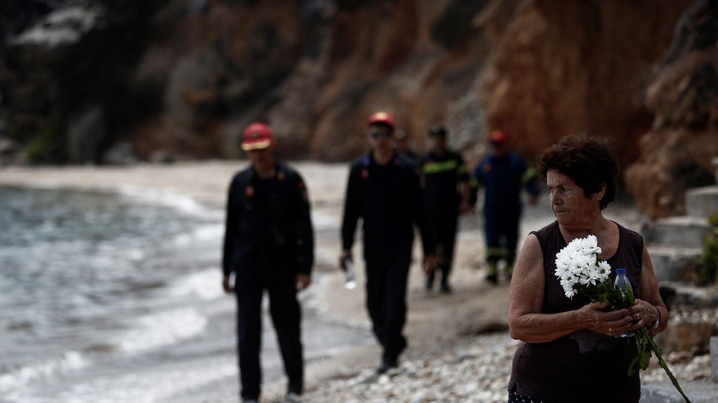 Varios bomberos buscan a desaparecidos mientras una mujer sostiene un ramo de flores tras el incendio forestal de Mati