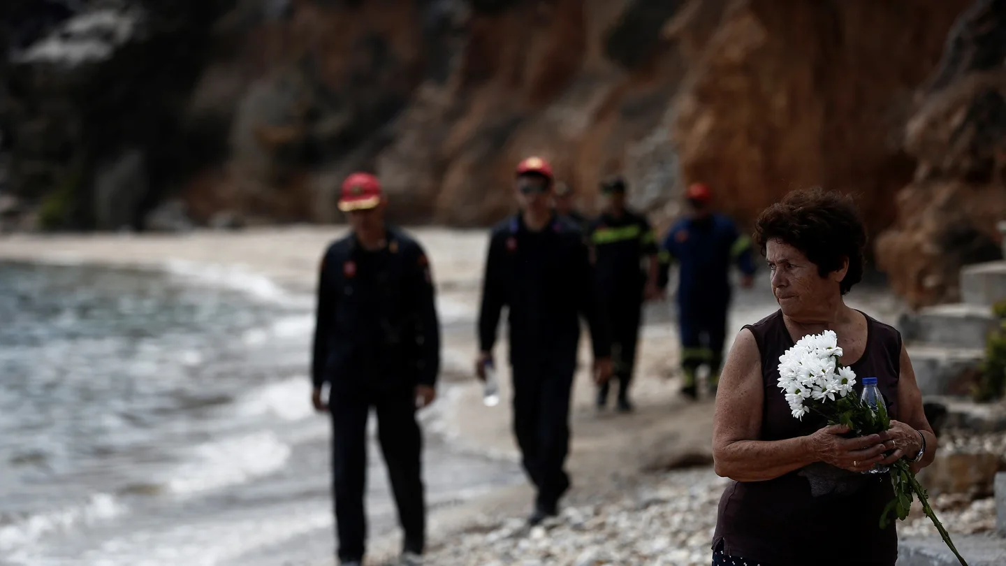 Varios bomberos buscan a desaparecidos mientras una mujer sostiene un ramo de flores tras el incendio forestal de Mati Varios bomberos buscan a desaparecidos mientras una mujer sostiene un ramo de flores tras el incendio forestal de Mati
