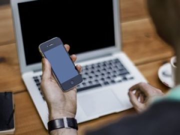 Un hombre sostiene un smartphone frente a un ordenador portátil. Un hombre sostiene un smartphone frente a un ordenador portátil.