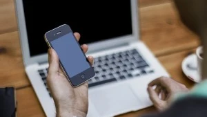 Un hombre sostiene un smartphone frente a un ordenador portátil.