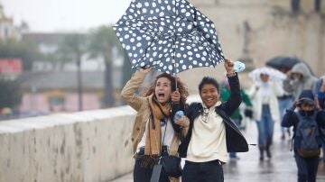 Unas turistas intentan protegerse del viento y de la lluvia con un paraguas mientras caminan por el puente romano de Córdoba