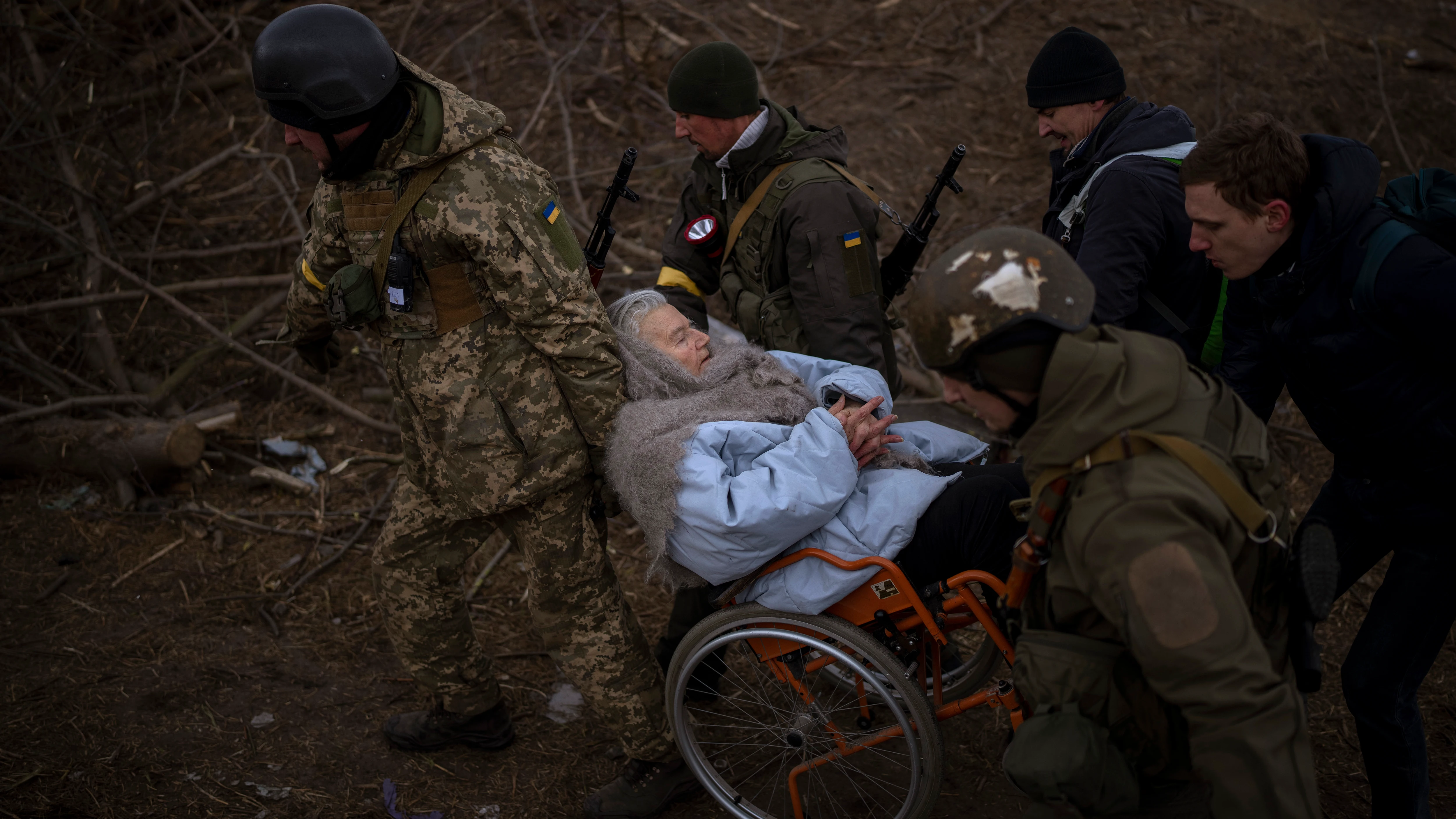 Soldados y milicianos ucranianos cargan a una mujer en silla de ruedas mientras la artillería resuena cerca, mientras la gente huye de Irpin en las afueras de Kiev, Ucrania. 