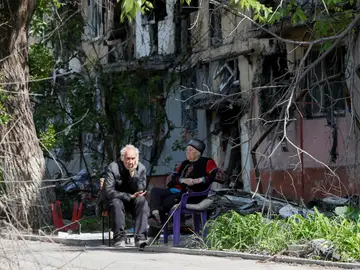 Dos personas sentadas frente a un edificio en ruinas por los ataques rusos en Mariúpol