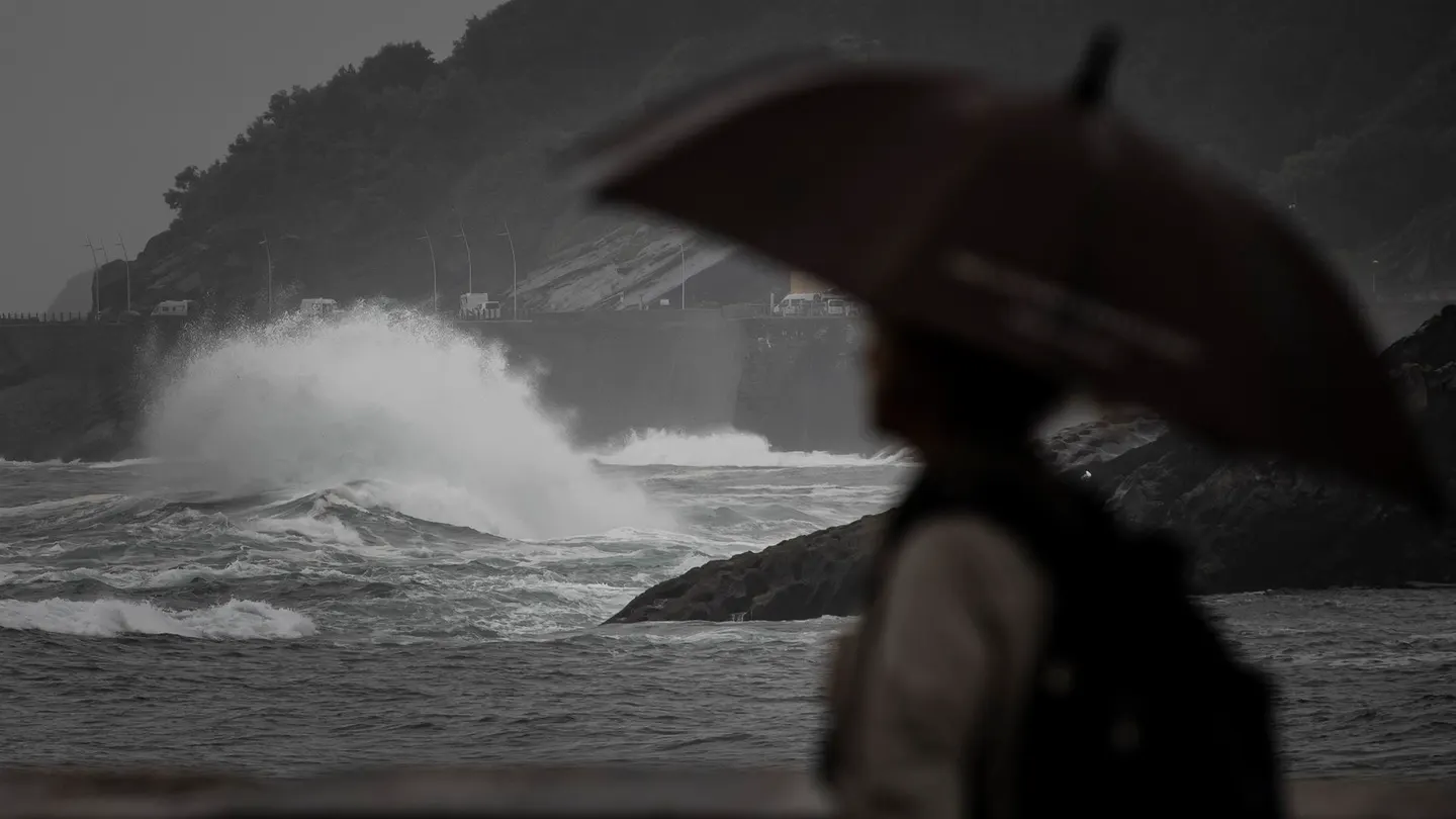Una persona sostiene un parahuas ante un mar embravecido Una persona sostiene un parahuas ante un mar embravecido