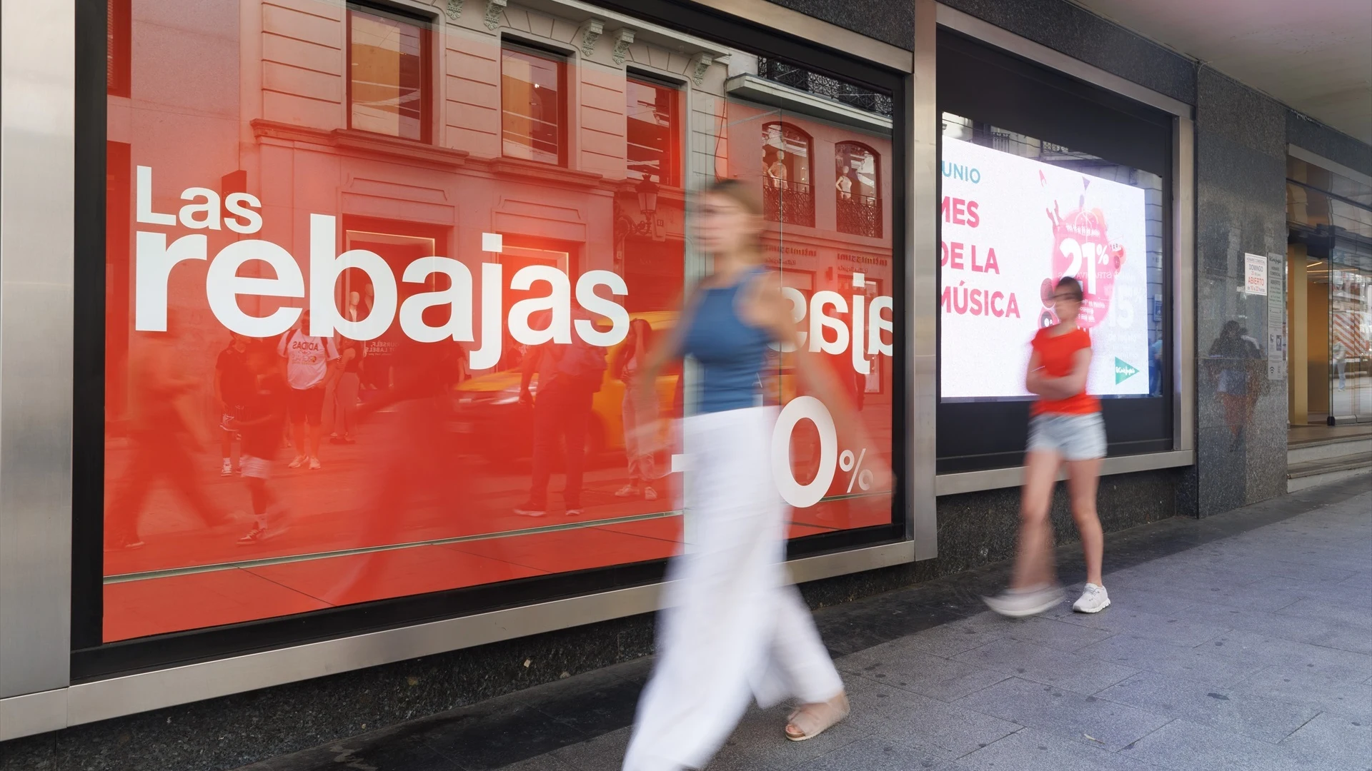 Varias personas pasan al lado de un cartel de rebajas en un escaparate el día que comienzan las rebajas de verano 2023