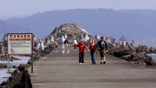 Una familia, caminando ante un cartel en el que se puede leer 'Peligro: olas extremadamente peligrosas' en Crescent City Una familia, caminando ante un cartel en el que se puede leer 'Peligro: olas extremadamente peligrosas' en Crescent City