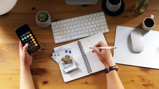 Planificación financiera post verano: recupera tu economía después de las vacaciones. Una persona hace cuentas en una libreta, mientras usa la calculadora y agrupa billetes y monedas de euro en su escritorio.