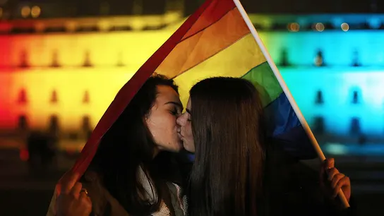 Imagen de archivo de una pareja de mujeres besándose con una bandera arcoíris. Imagen de archivo de una pareja de mujeres besándose con una bandera arcoíris.