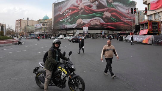 Un hombre con su motocicleta frente a una enorme pancarta que muestra manos sosteniendo banderas iran&iacute;es como se&ntilde;al de patriotismo, en Teher&aacute;n, este mi&eacute;rcoles.