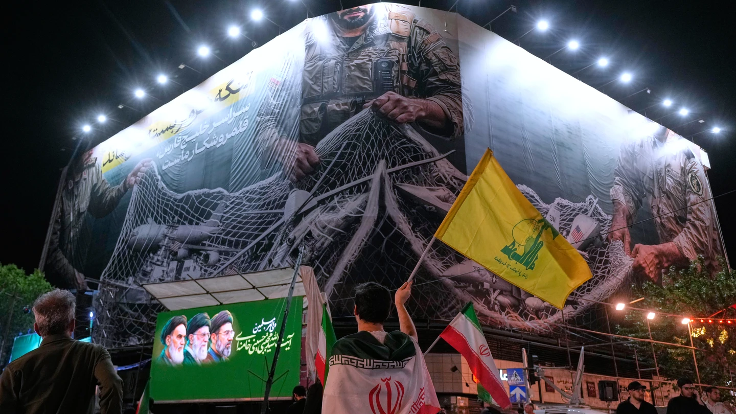 Un manifestante iraní ondea una bandera de Hizbulá bajo una valla que muestra aviones estadounidenses cayendo en la red de pesca de las fuerzas armadas iraníes Un manifestante iraní ondea una bandera de Hizbulá bajo una valla que muestra aviones estadounidenses cayendo en la red de pesca de las fuerzas armadas iraníes