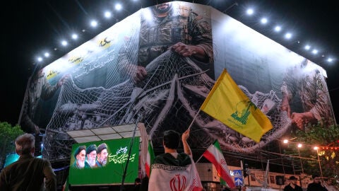 Un manifestante iran&iacute; ondea una bandera de Hizbul&aacute; bajo una valla que muestra aviones estadounidenses cayendo en la red de pesca de las fuerzas armadas iran&iacute;es