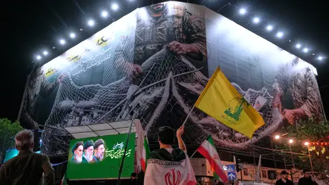 Un manifestante iraní ondea una bandera de Hizbulá bajo una valla que muestra aviones estadounidenses cayendo en la red de pesca de las fuerzas armadas iraníes Un manifestante iraní ondea una bandera de Hizbulá bajo una valla que muestra aviones estadounidenses cayendo en la red de pesca de las fuerzas armadas iraníes