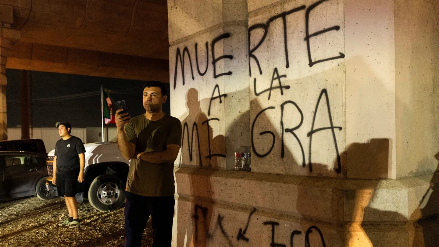 Varias personas cerca de una columna con el mensaje "Muerte a la Migra" (que significa "Muerte al ICE") mientras observan a manifestantes en el lugar donde un agente del ICE disparó a una persona en Austin Varias personas cerca de una columna con el mensaje "Muerte a la Migra" (que significa "Muerte al ICE") mientras observan a manifestantes en el lugar donde un agente del ICE disparó a una persona en Austin