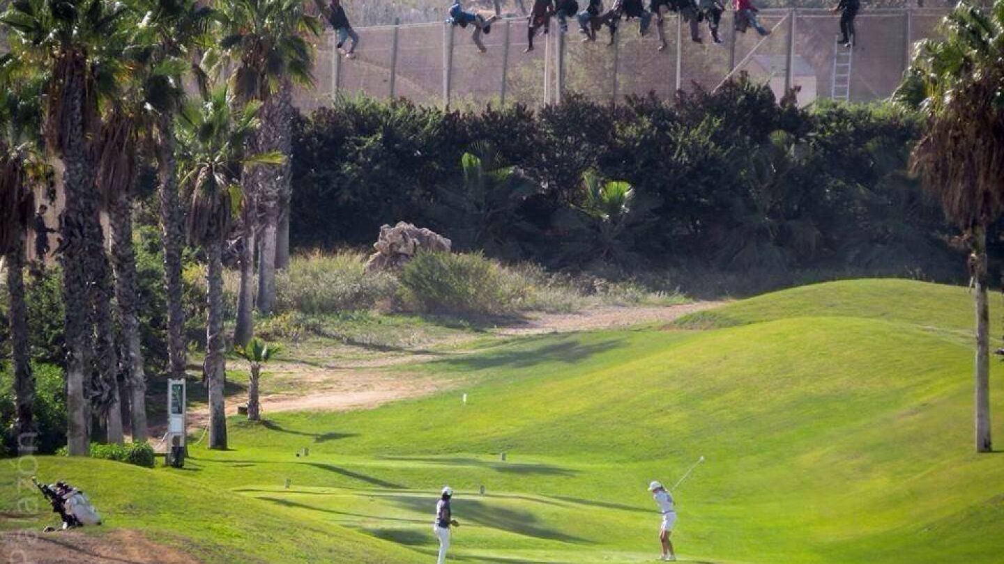 Inmigrantes en la valla de Melilla mientras varias personas juegan al golf