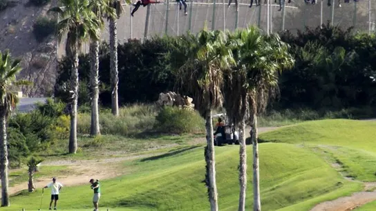 Inmigrantes encaramados en la valla de Melilla mientras varias personas juegan al golf Inmigrantes encaramados en la valla de Melilla mientras varias personas juegan al golf