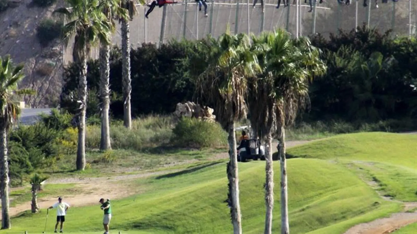 Inmigrantes encaramados en la valla de Melilla mientras varias personas juegan al golf Inmigrantes encaramados en la valla de Melilla mientras varias personas juegan al golf