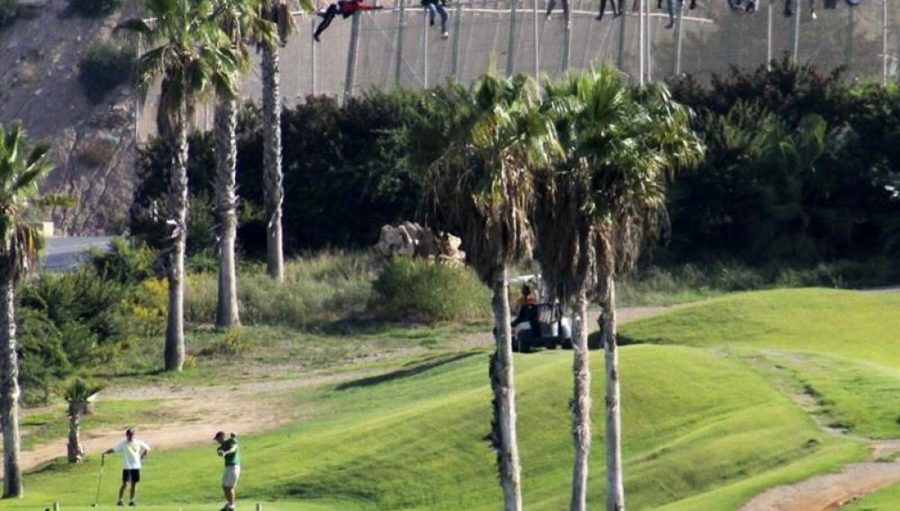 Inmigrantes encaramados en la valla de Melilla mientras varias personas juegan al golf