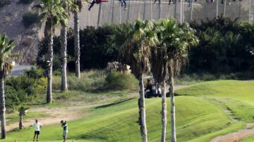 Inmigrantes encaramados en la valla de Melilla mientras varias personas juegan al golf