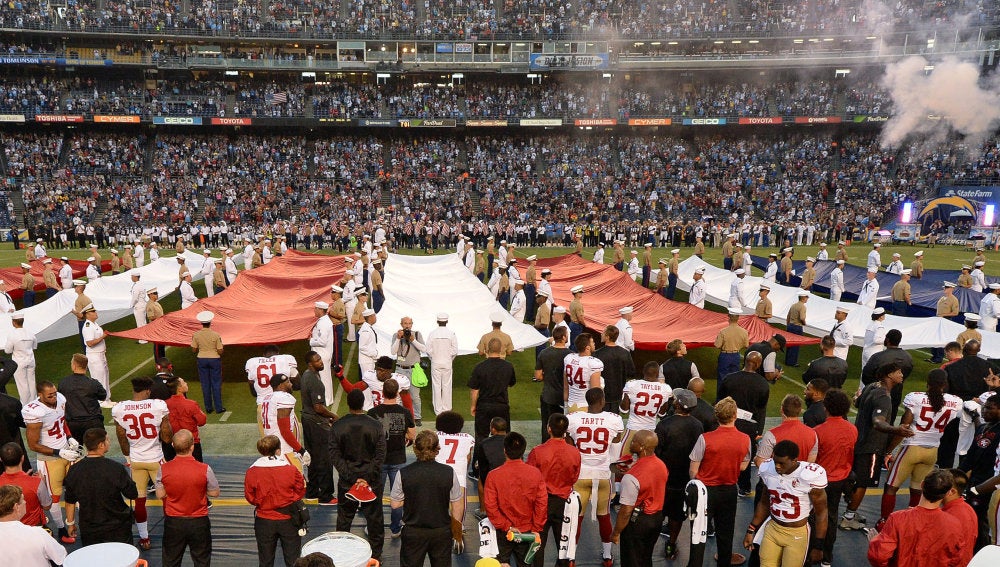 Colin Kaepernick, en la parte inferior de la fotografía (lleva el dorsal 7), de rodillas mientras suena el himno de Estados Unidos