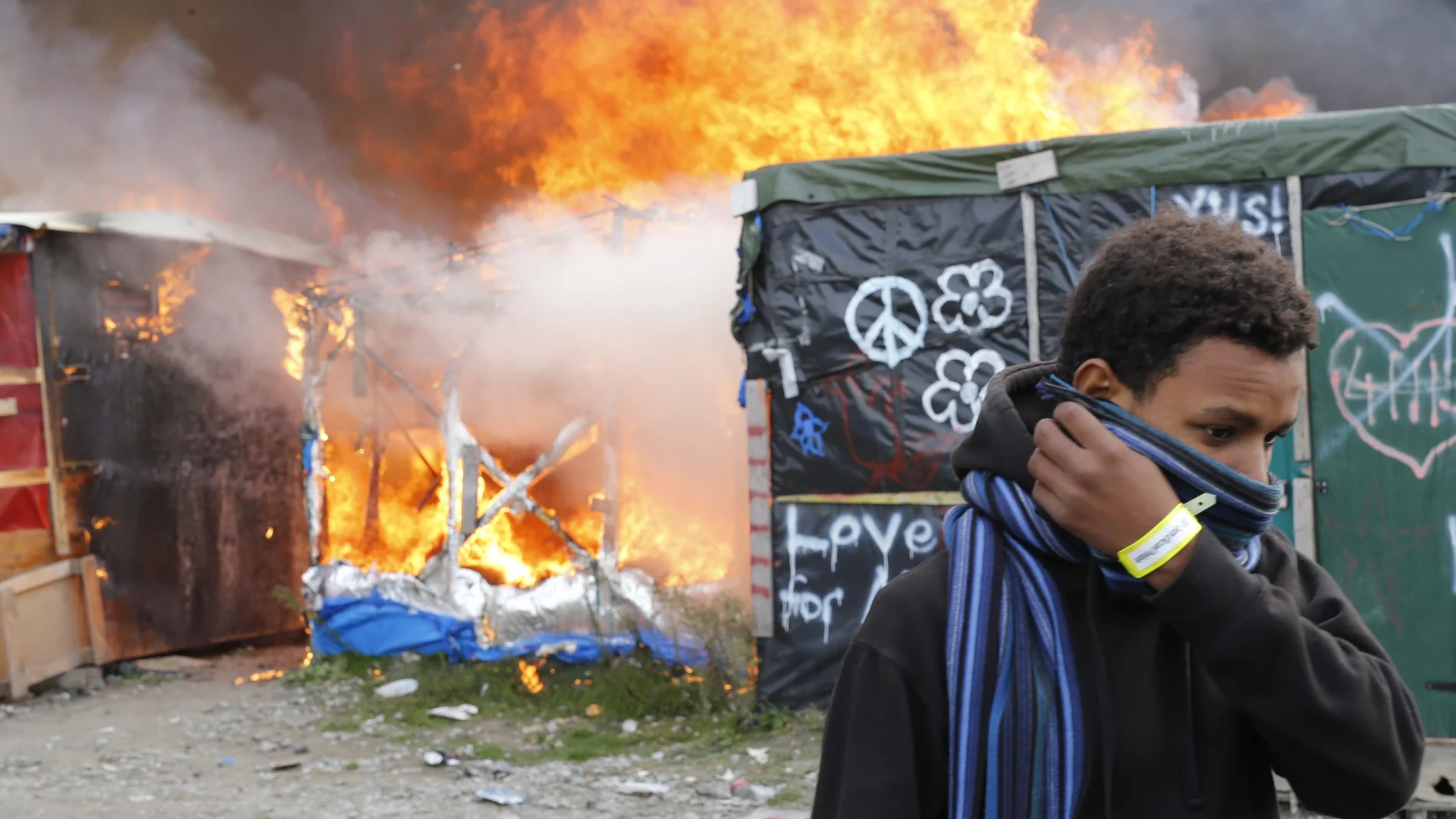 Un migrante se protege el rostro al pasar junto a chabolas en llamas durante el desmantelamiento del campamento conocido como la 'Jungla' de Calais 
