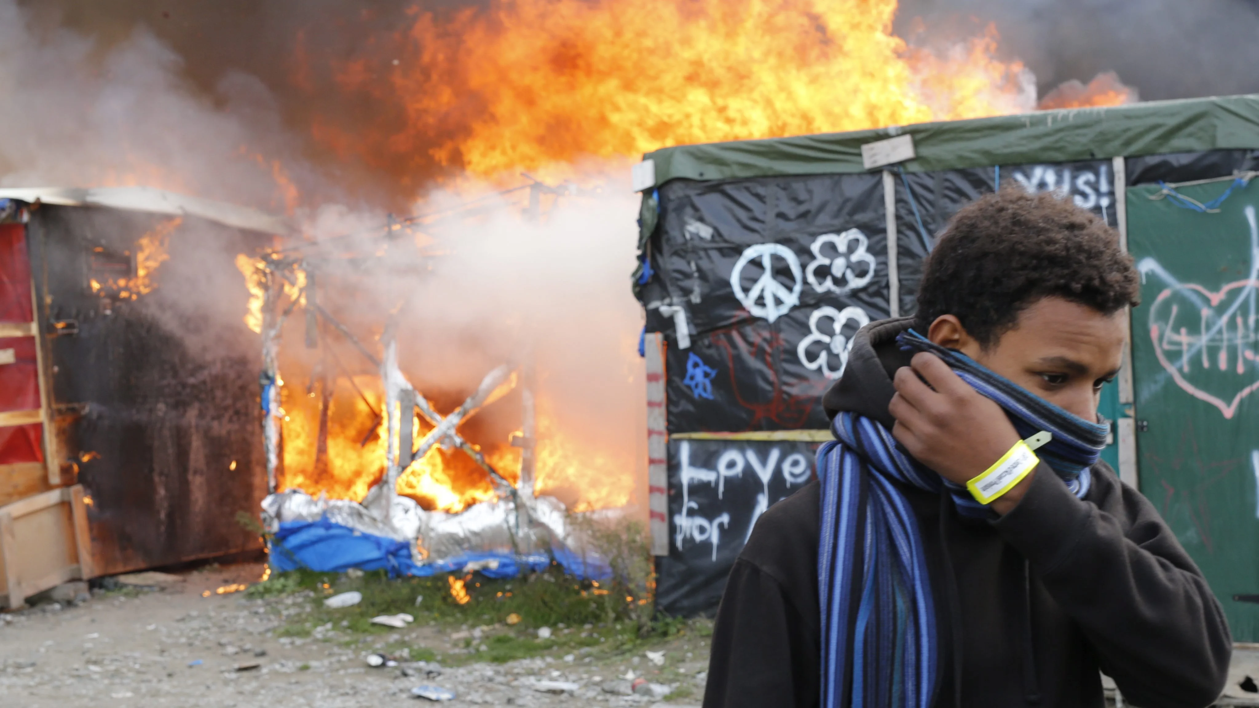 Un migrante se protege el rostro al pasar junto a chabolas en llamas durante el desmantelamiento del campamento conocido como la 'Jungla' de Calais 