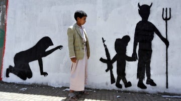 Un niño yemení observa el grafiti de la pared en la que se muestra a un niño soldado caminando de la mano de un demonio