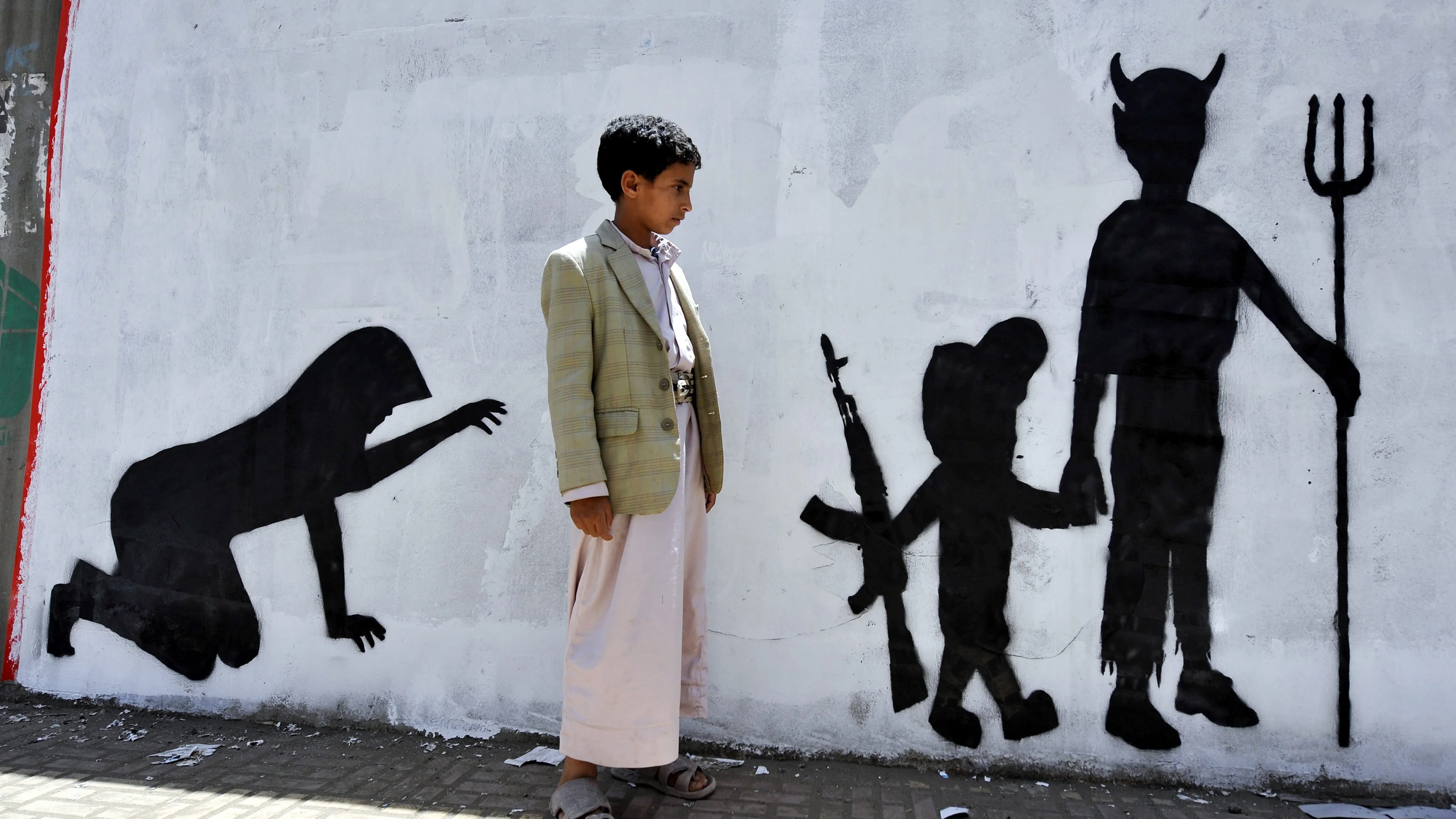 Un niño yemení observa el grafiti de la pared en la que se muestra a un niño soldado caminando de la mano de un demonio