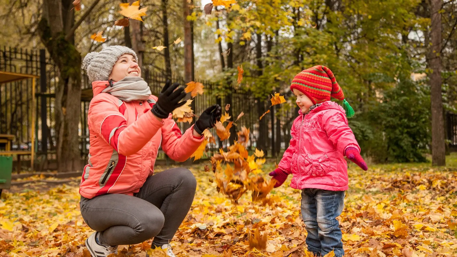 Fotografía otoñal de una mujer y un niño, jugando con las hojas caídas de los árboles, en un parque.