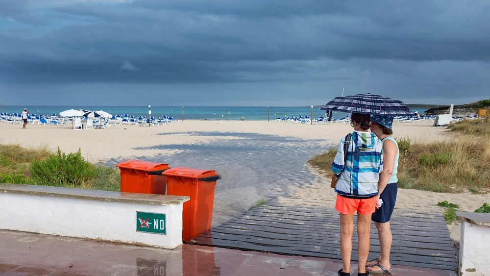 Dos mujeres se resguardan de la lluvia bajo un paraguas en una playa de Menorca