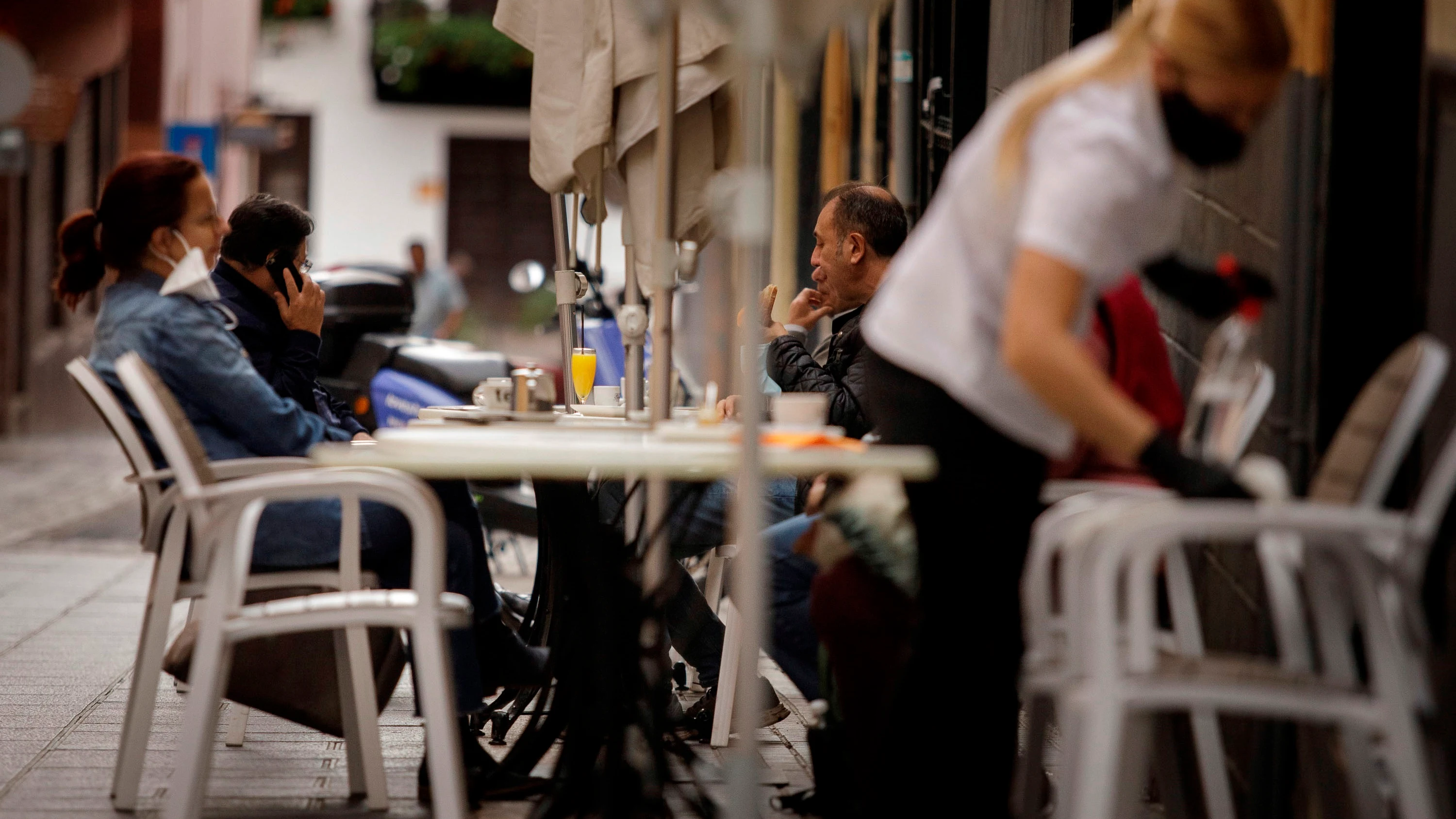 Una camarera de una cafetería de Santa Cruz de Tenerife limpia una mesa mientras varios clientes consumen en la terraza