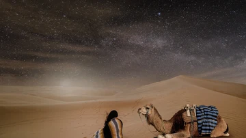 Una mujer observa el cielo nocturno en un desierto junto a su camello