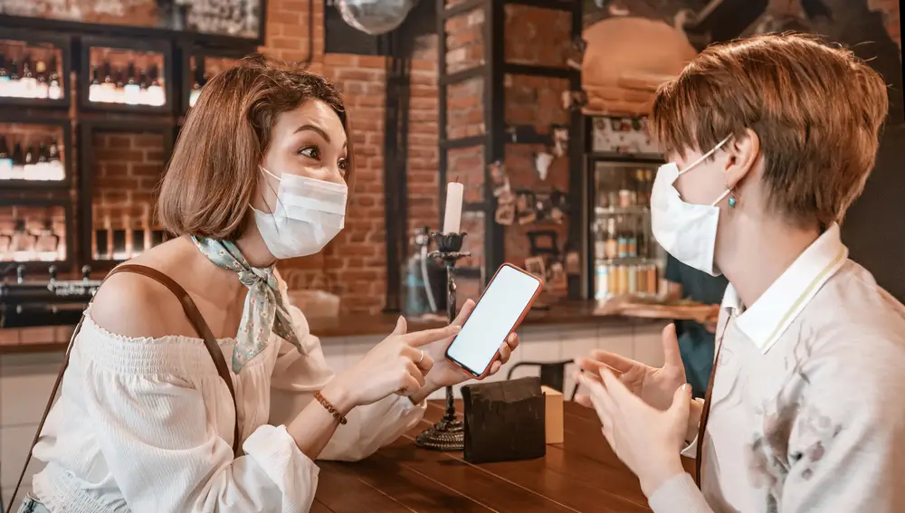 Dos jóvenes con mascarilla conversan en un bar mientras una sostiene un móvil