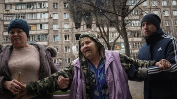 Personas ayudan a una anciana a caminar en una calle con un edificio de apartamentos alcanzado por los bombardeos de fondo en Mariupol