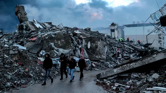 Gente caminando junto a los escombros de un edificio colapsado en Hatay. Gente caminando junto a los escombros de un edificio colapsado en Hatay.