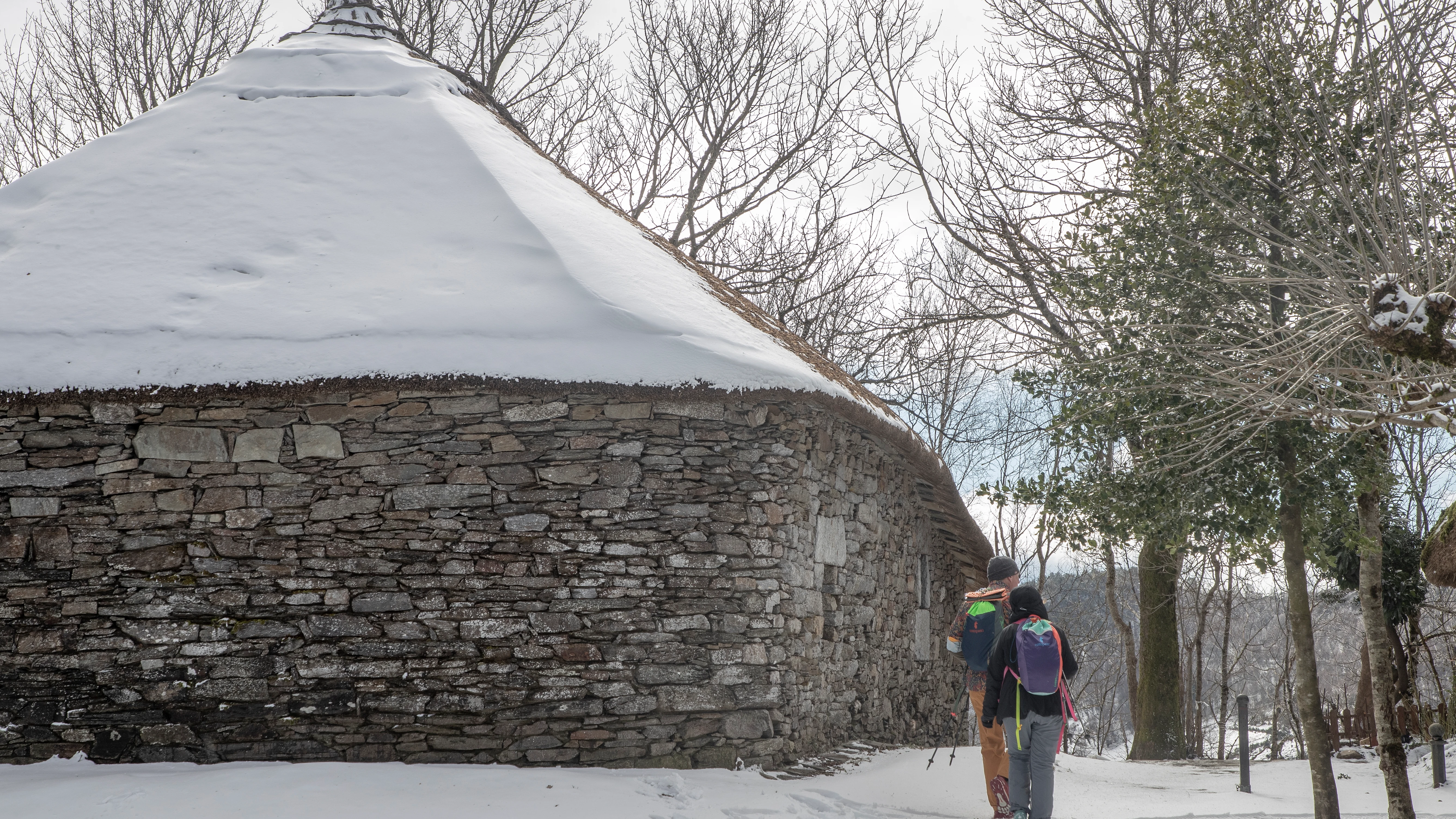 Dos peregrinos pasean cerca de una palloza cubierta por la nieve en el pueblo de O Cebreiro, Lugo