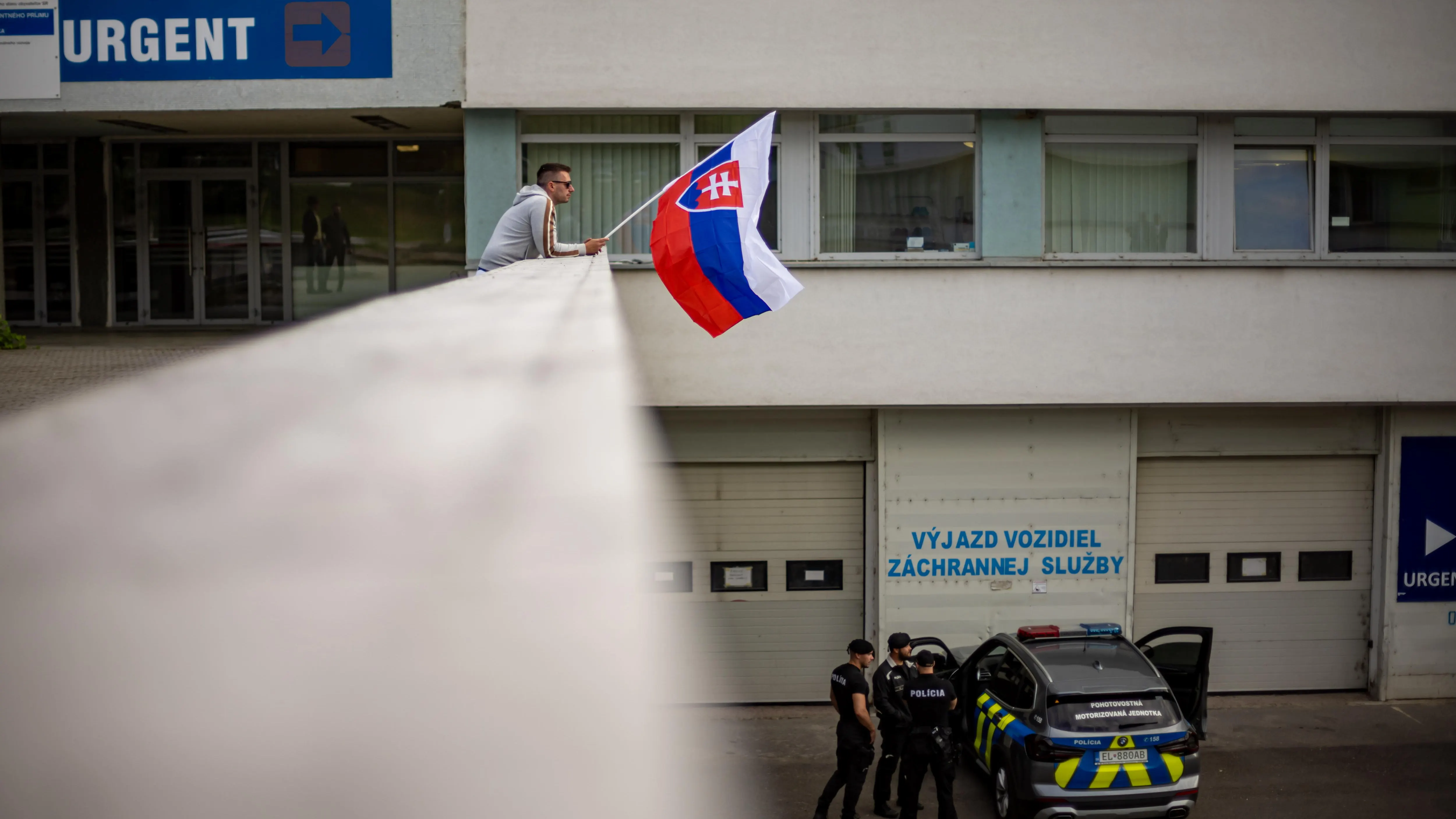 Un hombre ondea la bandera nacional eslovaca mientras un coche de policía está estacionado frente al Hospital Universitario F. D. Roosevelt, donde se encuentra el primer ministro eslovaco, Robert Fico.