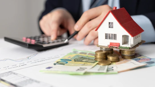 El euríbor es el indicador de referencia para las hipotecas. La figura de una casa se sostiene sobre monedas y billetes de euro mientras una persona hace cálculos con una calculadora.