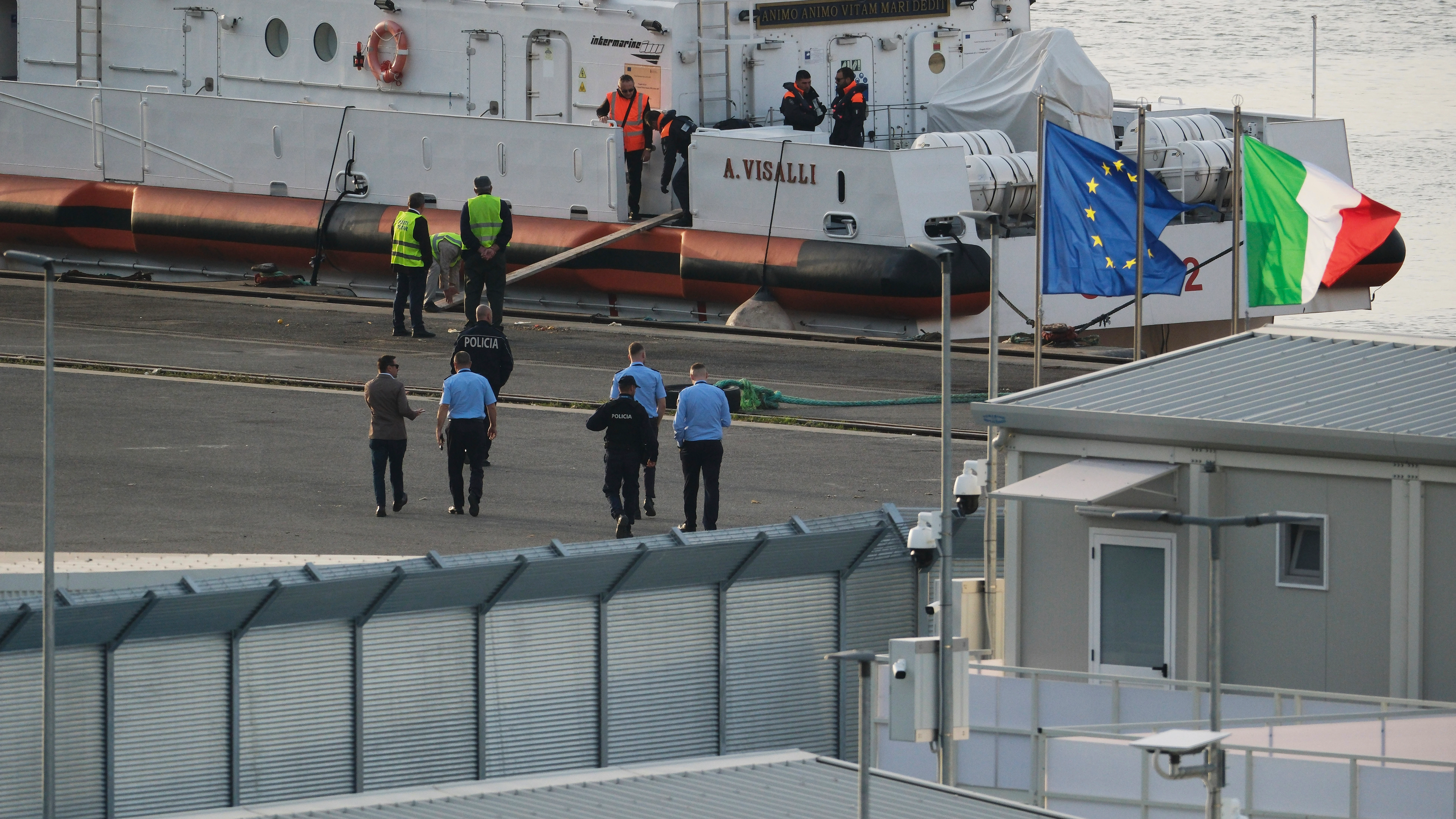 Un funcionario de seguridad camina frente a un barco de la guardia costera italiana que se prepara para partir del puerto de Shengjin