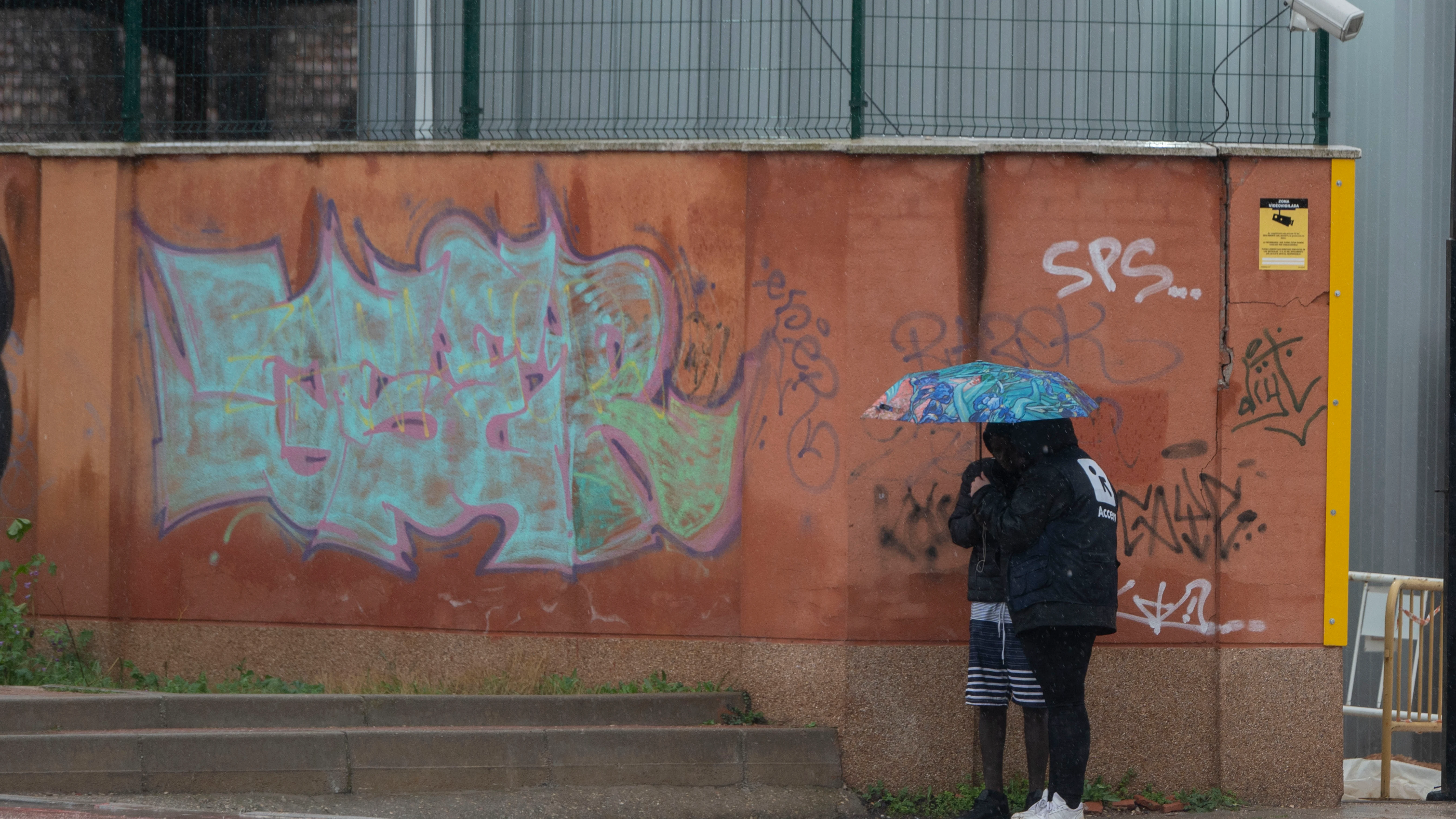 Dos migrantes se protegen de la lluvia con un paraguas en las inmediaciones del centro de Emergencia, Acogida y Derivación de Alcalá de Henares