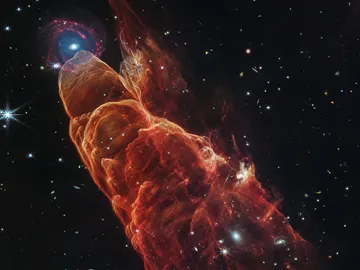 La nube de forma cónica de color rojo anaranjado corresponde a Herbig-Haro 40/50. En el extremo superior izquierdo hay una galaxia de fondo en forma de espiral con un centro azul. La nube de forma cónica de color rojo anaranjado corresponde a Herbig-Haro 40/50. En el extremo superior izquierdo hay una galaxia de fondo en forma de espiral con un centro azul.