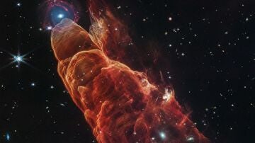 La nube de forma cónica de color rojo anaranjado corresponde a Herbig-Haro 40/50. En el extremo superior izquierdo hay una galaxia de fondo en forma de espiral con un centro azul.