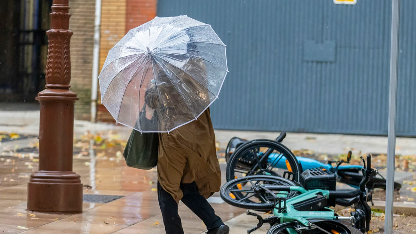 Una mujer con paraguas pasa ante unas bicicletas caídas en Huelva Activado el aviso rojo por lluvias en Huelva y enviado mensaje de alerta a los móviles