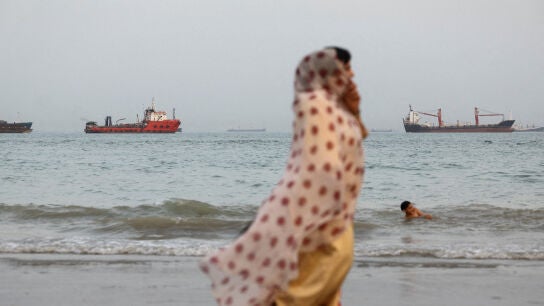 Personas caminan por la playa, con embarcaciones visibles en el estrecho de Ormuz cerca de la playa de Bandar Abbas, Ir&aacute;n