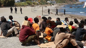Migrantes esperan en una playa bajo control policial en Ceuta, tratando de protegerse del intenso sol